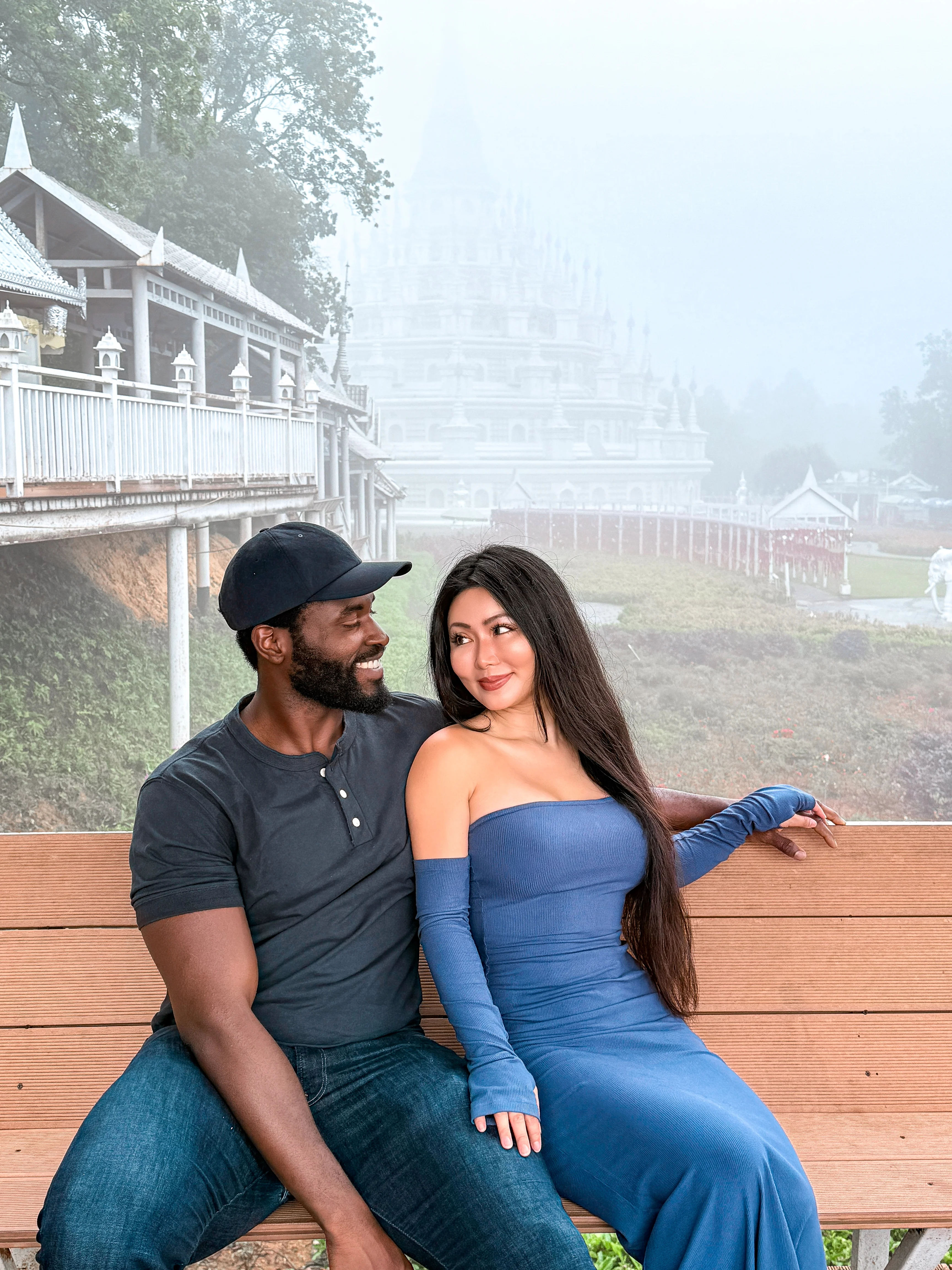 Theophilus and his wife seated close together on a wooden bench, smiling at each other, a white temple rising through mist behind them