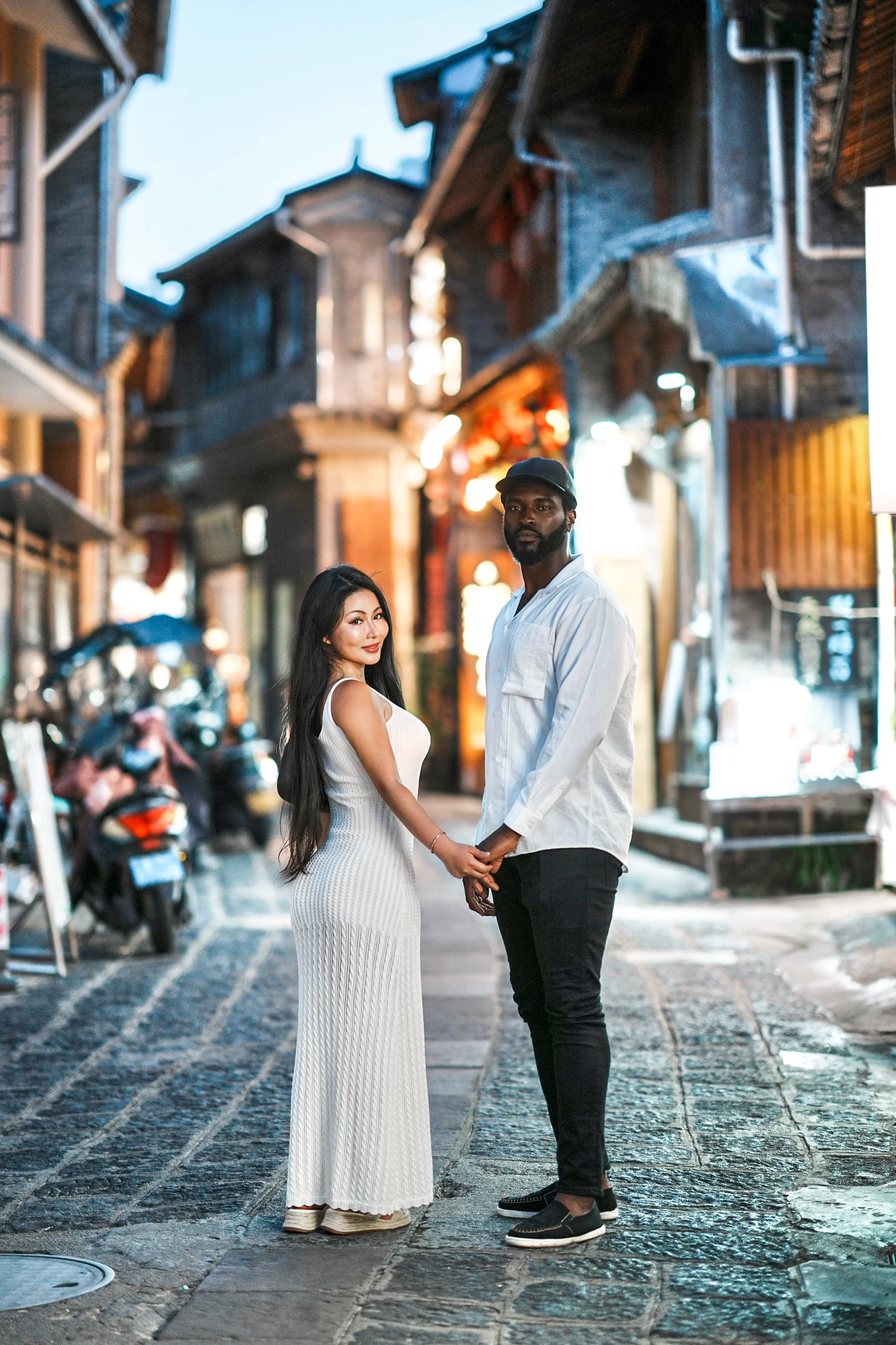 Theophilus and his wife holding hands in a lantern-lit old-town street at dusk; she glances back over her shoulder toward the camera